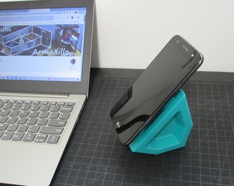 Puede incluir: Un soporte para teléfono impreso en 3D de color turquesa con un teléfono inteligente negro apoyado en la parte superior. El teléfono está ligeramente inclinado hacia arriba. Se puede ver un ordenador portátil en el fondo.