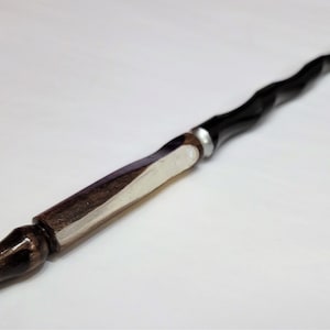 Puede incluir: Una varita de madera negra y marrón con un acento plateado. La varita está retorcida y tiene una punta puntiaguda.