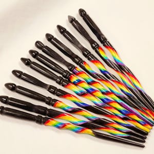 May include: A set of 12 black wooden wands with rainbow stripes. The wands are arranged in a fan shape, with the tips pointing towards the top of the image.