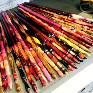 May include: A collection of handcrafted wooden wands in various colors, including red, pink, yellow, and black. Each wand has a unique twisted design and is displayed on a gray surface. The wands are from Orchard Works.