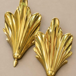 May include: A pair of polished brass wall sconces featuring a fan-shaped design with vertical fluted ridges. Each piece has a pointed finial at the base and a symmetrical, leaf-like silhouette with a reflective, metallic gold-toned finish.