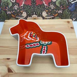 Puede incluir: Un cuenco de cerámica con forma de caballo de Dalecarlia, con un interior naranja vibrante y bordes exteriores blancos. El fondo del cuenco presenta un patrón decorativo pintado en verde, rojo y blanco, imitando el arnés tradicional de un caballo.