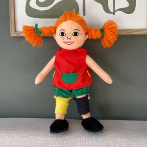 May include: A plush doll with orange yarn pigtails, freckles, and a red top with a green pocket. The doll wears green shorts, mismatched yellow and black leg warmers, and black shoes. The doll is a children's toy.