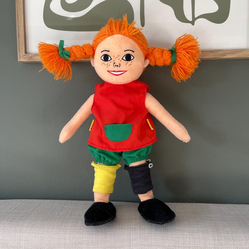 May include: A plush doll with orange yarn pigtails, freckles, and a red top with a green pocket. The doll wears green shorts, mismatched yellow and black leg warmers, and black shoes. The doll is a children's toy.