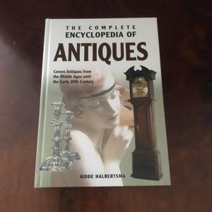 Peut inclure: Un livre à couverture rigide intitulé "THE COMPLETE ENCYCLOPEDIA OF ANTIQUES" avec une image de femme, une horloge de grand-père et des chandeliers en argent. Le livre traite des antiquités du Moyen Âge au début du XXe siècle.