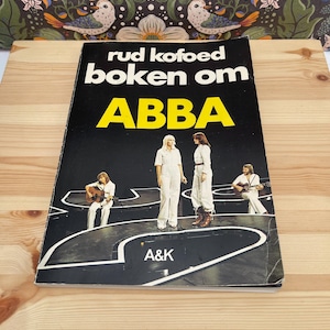 Op de afbeelding: Een paperback boek getiteld "rud kofoed boken om ABBA" met een zwarte omslag met een foto van de vier leden van de band ABBA in witte outfits op een podium. De tekst "ABBA" is gedrukt in grote gele letters en "A&K" staat onderaan.