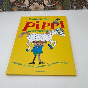 Può includere: Un libro per bambini giallo intitolato "Känner Du Pippi Långstrump?" presenta un'illustrazione a cartoni animati di una ragazza che tiene in mano un cavallo bianco con macchie blu. La copertina del libro include anche testo in rosso e verde.