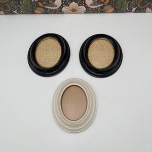 May include: Three oval picture frames. Two frames are black with a light brown interior, and one frame is off-white with a light brown interior. The frames are arranged on a white surface.