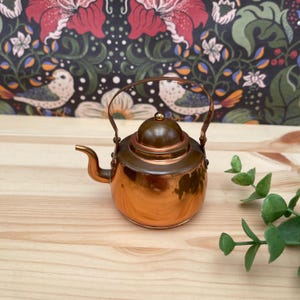 May include: A miniature copper-coloured teapot with a rounded body, a curved spout, and a thin metal handle. The lid features a small gold-toned knob at the centre. The teapot has a polished, reflective surface that catches light, showing a warm metallic tone.