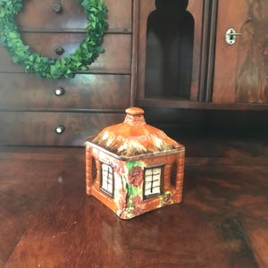 May include: A small ceramic cottage-shaped sugar bowl with a thatched roof and floral details. The bowl is brown with green and red accents.