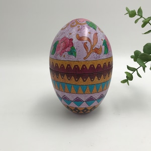 Op de afbeelding: Een eivormige decoratieve doos met een scharnierend deksel. De doos is beschilderd met een bloemmotief in roze, goud en blauwgroen. De basis van de doos heeft een geometrisch patroon. De doos is waarschijnlijk van metaal.