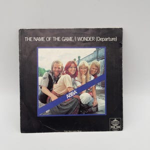 Könnte beinhalten: Vintage ABBA Schallplattenhülle mit dem Text "THE NAME OF THE GAME/I WONDER (Departure)". Das Cover ist schwarz. Im blauen Rahmen befindet sich ein Foto der Band. Das ABBA-Logo ist sichtbar. Die Hülle ist leicht abgenutzt.