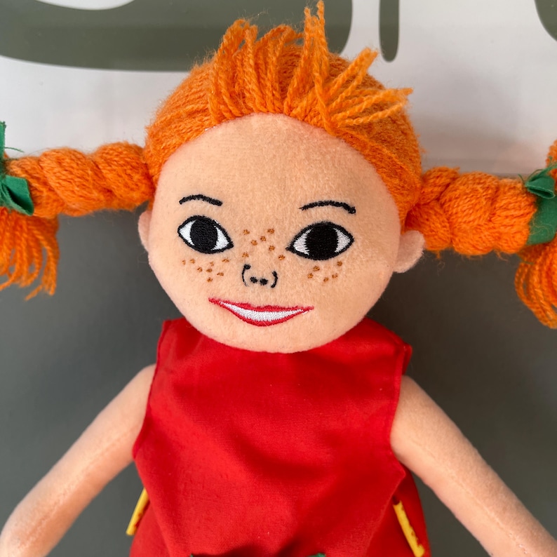 May include: A plush doll with orange yarn pigtails and hair, freckles, and a red dress. The doll has a smiling expression and green bows at the base of the pigtails. The doll's arms are visible.