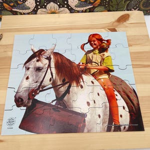 May include: A jigsaw puzzle featuring a red-haired child with braided hair sitting on a white horse with brown spots. The child wears a yellow shirt and orange stockings. A small monkey sits on the child's shoulder. The puzzle includes the text Pippi L&aring;ngstrump.