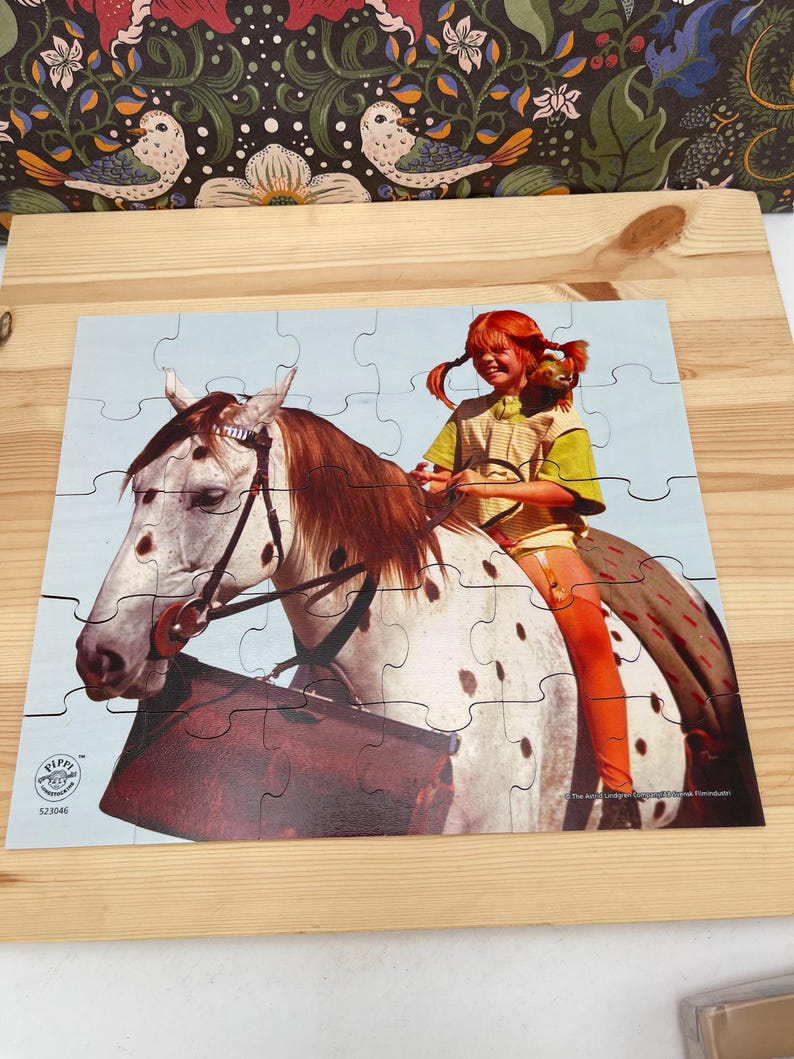 May include: A jigsaw puzzle featuring a red-haired child with braided hair sitting on a white horse with brown spots. The child wears a yellow shirt and orange stockings. A small monkey sits on the child's shoulder. The puzzle includes the text Pippi L&aring;ngstrump.