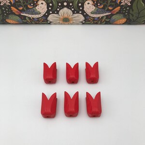 May include: Six small, hand-carved wooden tulip shapes painted a vibrant red. The tulips are arranged in two rows of three against a white background. The top of the image features a decorative floral pattern with birds.
