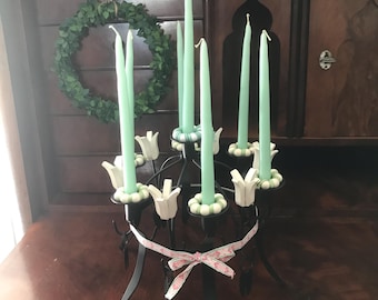 May include: A black metal candelabra with six arms, each with a white ceramic flower-shaped candle holder. The candelabra is decorated with a green and white ribbon and has six teal blue candles.