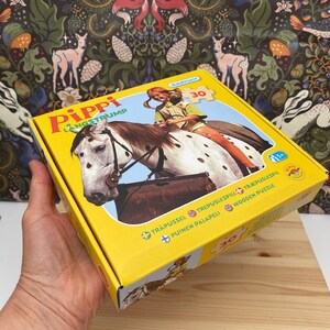 May include: A yellow cardboard box for a 30-piece wooden puzzle featuring Pippi Longstocking riding a spotted horse. The box displays the text Pippi Langstrump, Karnan, and Wooden Puzzle, with an illustration of the character and horse on the front.