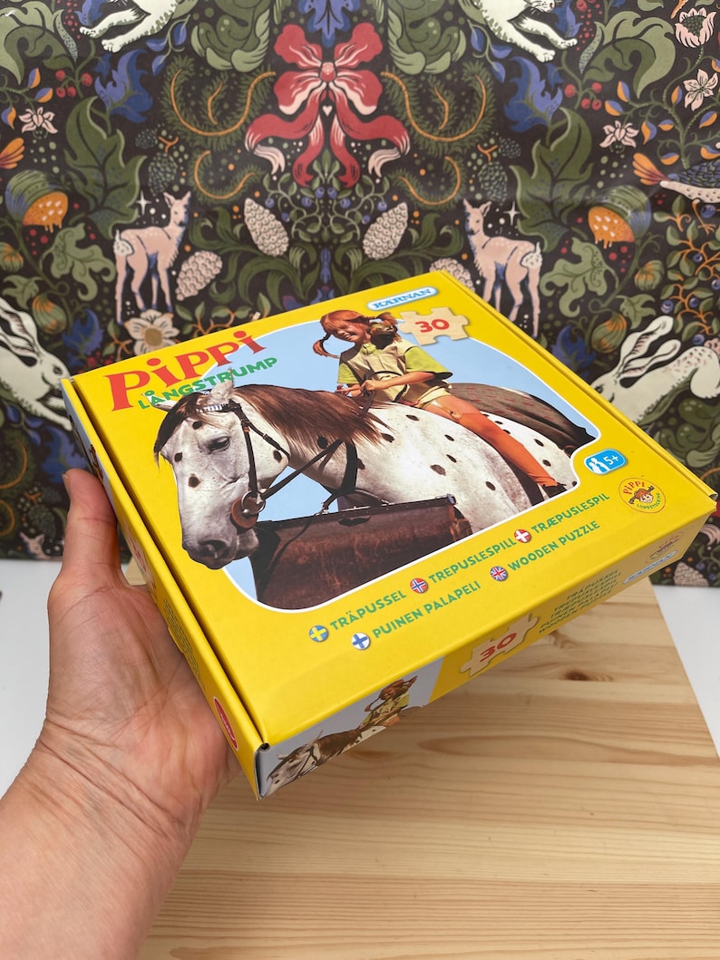 May include: A yellow cardboard box for a 30-piece wooden puzzle featuring Pippi Longstocking riding a spotted horse. The box displays the text Pippi Langstrump, Karnan, and Wooden Puzzle, with an illustration of the character and horse on the front.