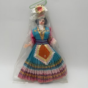 May include: A doll in traditional attire, packaged in a clear plastic bag. The doll wears a teal dress with a layered skirt, pink sleeves, and a decorative head covering. Gold accents and a white lace apron complete the outfit. A round pin is attached to the bag.