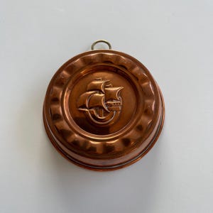 May include: A round copper mold featuring a raised embossed design of a sailing ship in the center. The mold has a scalloped edge and a small metal hanging loop attached to the top rim, providing a functional element for display or storage.
