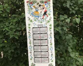 May include: A white linen calendar for the year 1999 with a floral border and a picture of a couple in traditional folk costumes dancing. The calendar includes months from January to December.