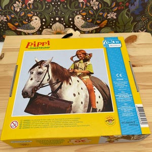 May include: A yellow rectangular box for a 30-piece wooden puzzle featuring Pippi Longstocking riding a spotted horse. The box displays the text Pippi L&aring;ngstrump, a 5+ age rating, and a 250 x 210 mm dimension label. The packaging includes safety warnings.