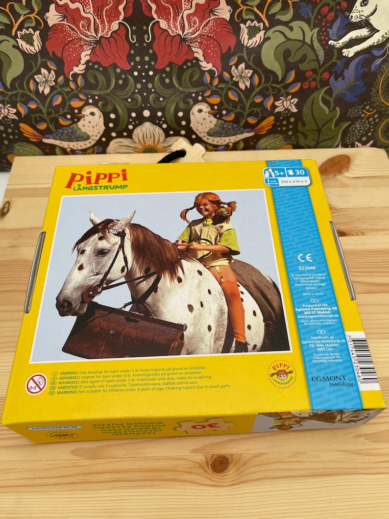 May include: A yellow rectangular box for a 30-piece wooden puzzle featuring Pippi Longstocking riding a spotted horse. The box displays the text Pippi L&aring;ngstrump, a 5+ age rating, and a 250 x 210 mm dimension label. The packaging includes safety warnings.