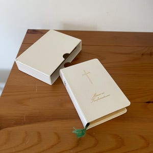 May include: A small, cream-colored book with a matching slipcase. The cover features a gold-colored cross and the text "Minne av Konfirmationen" in gold script. A thin green ribbon bookmark extends from the bottom edge of the book.