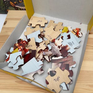 May include: A collection of wooden jigsaw puzzle pieces inside a yellow cardboard box. Several pieces are flipped over to reveal a light wood grain back, with the name Pippi handwritten in blue ink on some of the pieces. The front sides show colorful imagery.