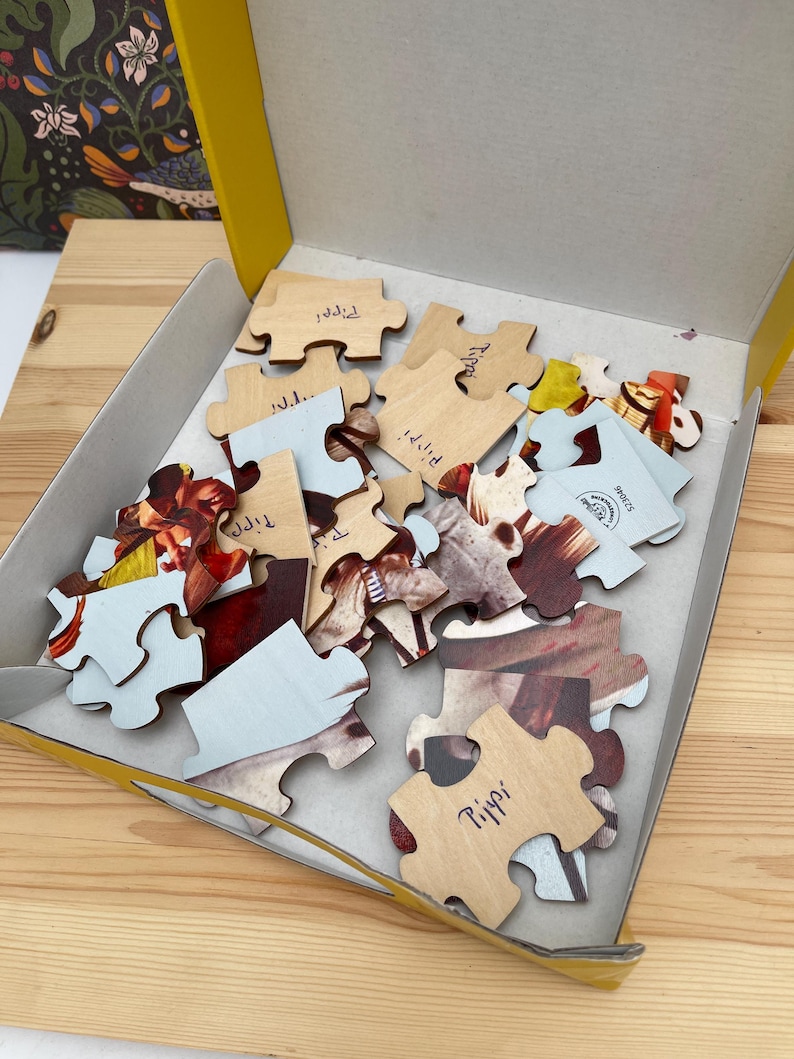 May include: A collection of wooden jigsaw puzzle pieces inside a yellow cardboard box. Several pieces are flipped over to reveal a light wood grain back, with the name Pippi handwritten in blue ink on some of the pieces. The front sides show colorful imagery.