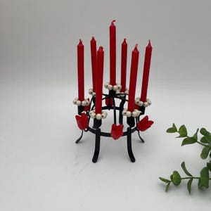 May include: A black metal candle holder with eight red candles. The candle holder features a circular design with white beads and red tulip accents. The candles are arranged in a circular pattern.