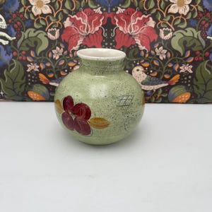 Könnte beinhalten: Kleine, runde, hellgrüne Keramikvase mit weißem Rand. Die Vase ist mit einer Ansammlung roter und gelber Blumendesigns verziert. Die Vase hat eine gesprenkelte Textur und ein einzigartiges, handgefertigtes Aussehen.
