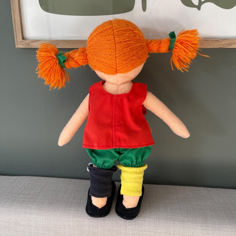 May include: A handmade doll with orange yarn pigtails tied with green ribbons. The doll wears a red top, green pants, and mismatched black and yellow shoes. The doll is a children's toy.
