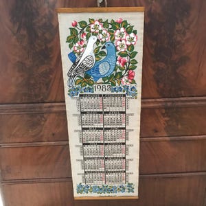 May include: A vintage 1983 calendar with a floral design featuring two birds. The calendar is printed on fabric and has a wooden frame. The calendar is in Swedish.