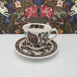 Può includere: Un set vintage di tazza e piattino da tè. La tazza e il piattino sono bianchi con un dettagliato motivo floreale e paisley nei toni del marrone, arancione e giallo. Il set è posto su una superficie bianca, con uno sfondo a fantasia.