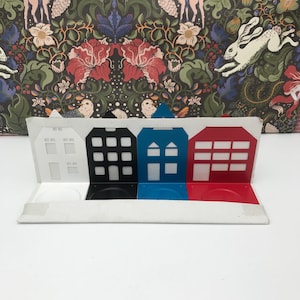Peut inclure: Un ensemble de quatre bougeoirs en métal en forme de maisons. Les maisons sont peintes en blanc, noir, bleu et rouge. Chaque maison a un petit bougeoir au fond.