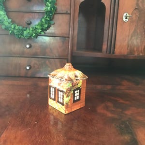 May include: A ceramic house-shaped container with a thatched roof and two windows. The container is painted with orange, brown, and green colours. The windows have white frames and black panes. The container is sitting on a wooden surface.