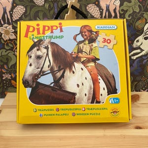 May include: A yellow cardboard box for a 30-piece wooden puzzle featuring Pippi Longstocking riding a spotted horse. The box displays the text Pippi Langstrump, K&auml;rnan, and includes icons for age 5 plus. The design shows a child with red braids.