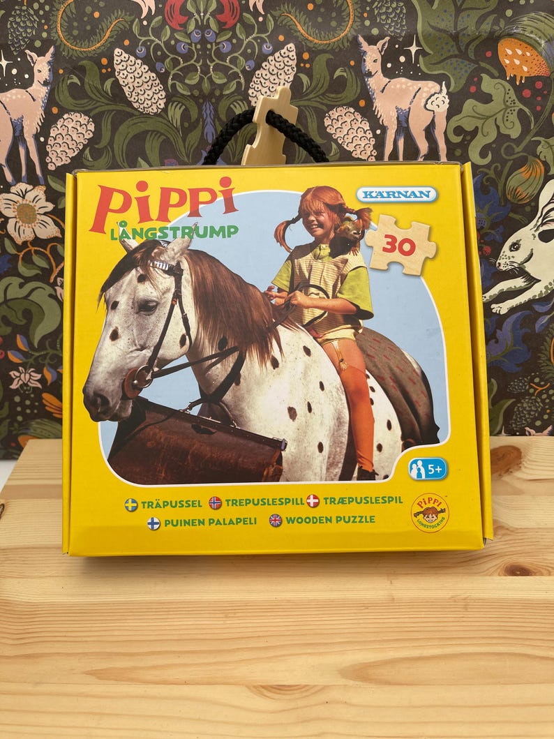 May include: A yellow cardboard box for a 30-piece wooden puzzle featuring Pippi Longstocking riding a spotted horse. The box displays the text Pippi Langstrump, K&auml;rnan, and includes icons for age 5 plus. The design shows a child with red braids.