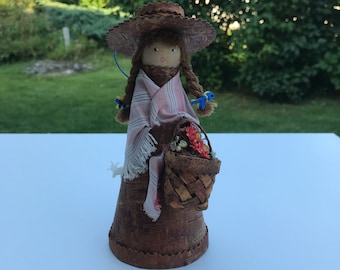 May include: A handmade wooden doll with a brown hat, pink and white striped dress, and a basket of flowers. The doll is made of birch bark and has a whimsical, folk art style.