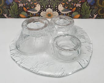 May include: Clear glass serving dish with three circular compartments and a textured, irregular base. The dish is designed to hold small items and has a unique, organic shape. The glass has a frosted appearance.