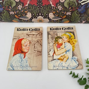 May include: Two vintage children's books titled "Kulla-Gulla" by Martha Sandwall-Bergström. One book features a girl in a red headscarf, the other shows a woman holding a baby. The books are set against a floral patterned background.