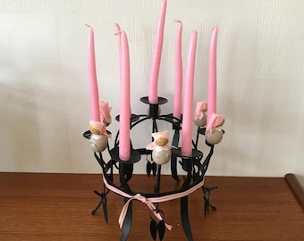 May include: A black metal candelabra with seven pink candles. The candelabra has a decorative design with small figures and a pink ribbon. The candles are long and slender, and the overall design is ornate.