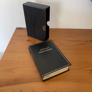 May include: A black book with gold lettering that reads Minne av konfirmationen, featuring a textured cover and a matching black slipcase. The book has cream-colored pages and a thin gold border on the front cover, presented as a set.