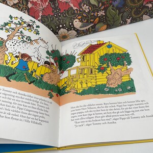 May include: An open book with colorful illustrations. One page shows a yellow house with a red roof and a tree with red apples. The opposite page has text in a foreign language. The book is open on a white surface.