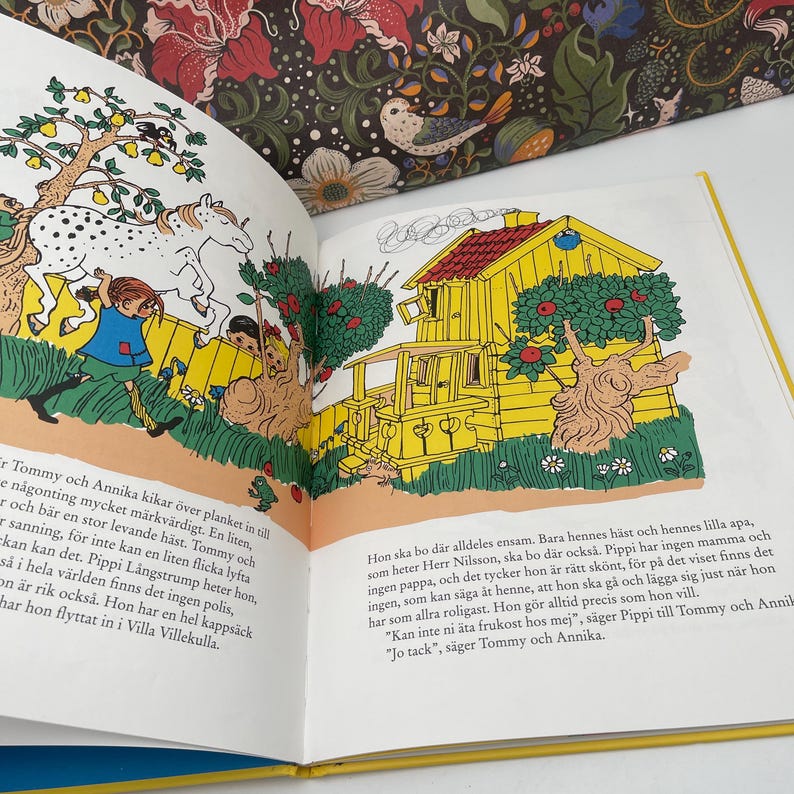 May include: An open book with colorful illustrations. One page shows a yellow house with a red roof and a tree with red apples. The opposite page has text in a foreign language. The book is open on a white surface.