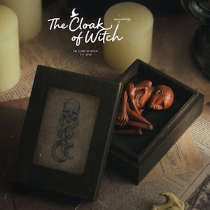 May include: A dark wooden box with a lid featuring a skull and snake illustration. Inside, a small, reddish-brown, emaciated humanoid figure is curled up. The text "The Cloak of Witch" and "EST 2022" is visible above the box, alongside a wand.
