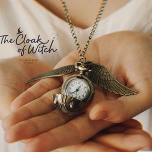 May include: A bronze-colored pocket watch necklace with wings, held in open hands. The watch face has white numbers and hands. The necklace is from "The Cloak of Witch".