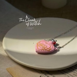 May include: A pink heart-shaped pendant necklace with the words "Love Potion" on the front. The necklace has a silver chain and is displayed on a white plate. The brand name "The Cloak of Witch" is visible.